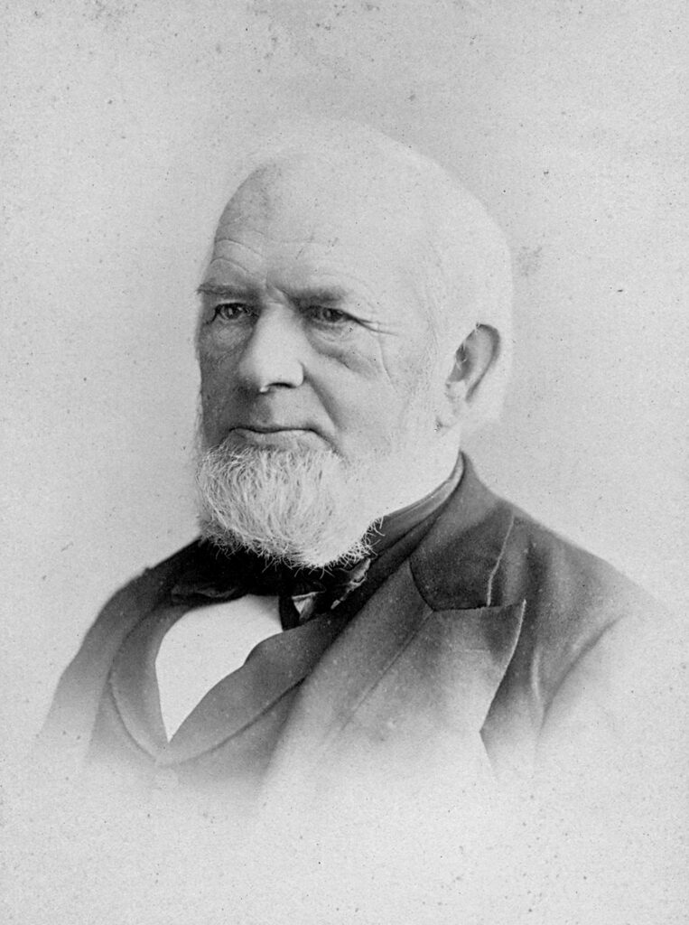Black and white portrait photograph shwoing the head and shoulders of an older white man with a full white beard.