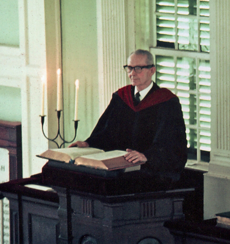 White man in a black preaching gown standing in the pulpit of the First Parish Meeting House.
