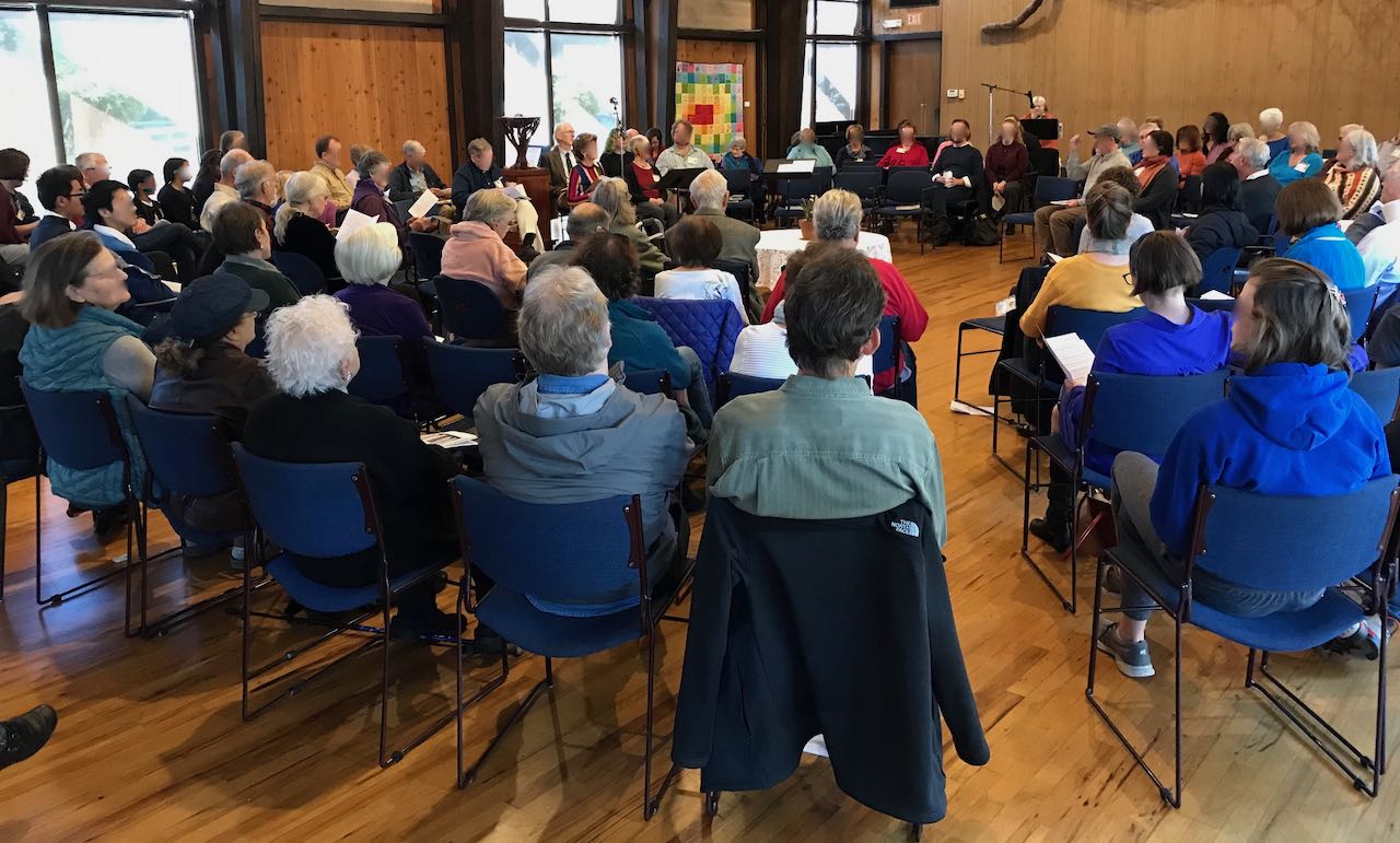 A worship service with people seated in concentric circles around an open central area, inside the Main Hall of the Unitarian Universalist Church of Palo Alto, which features large windows, wood floors and wood walls.