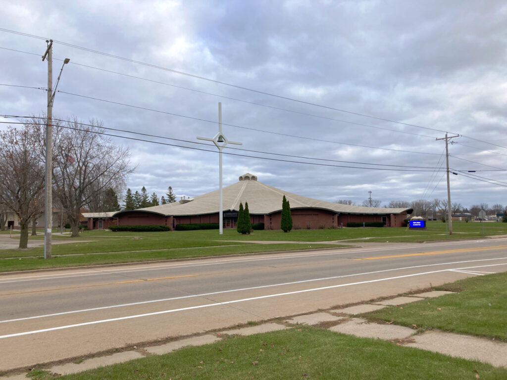Suburban streetscape, large one story building with low brick wall and gently sloping undulating roof. A large freestanding white cross is as tall as nearby utility poles