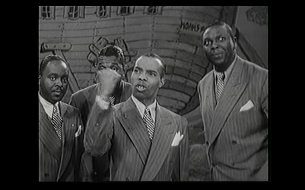 Black and white photo of four men wearing 1940s style suits and ties. One man stands in the center, and is the lead singer. The three others stand around and behind him.