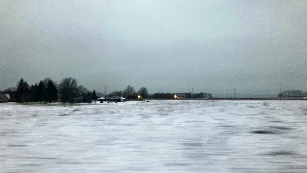 A cloudy sky over a snow covered field, with a line of trees in the distance, and through a break in the trees perhaps a sight of a distant lake.
