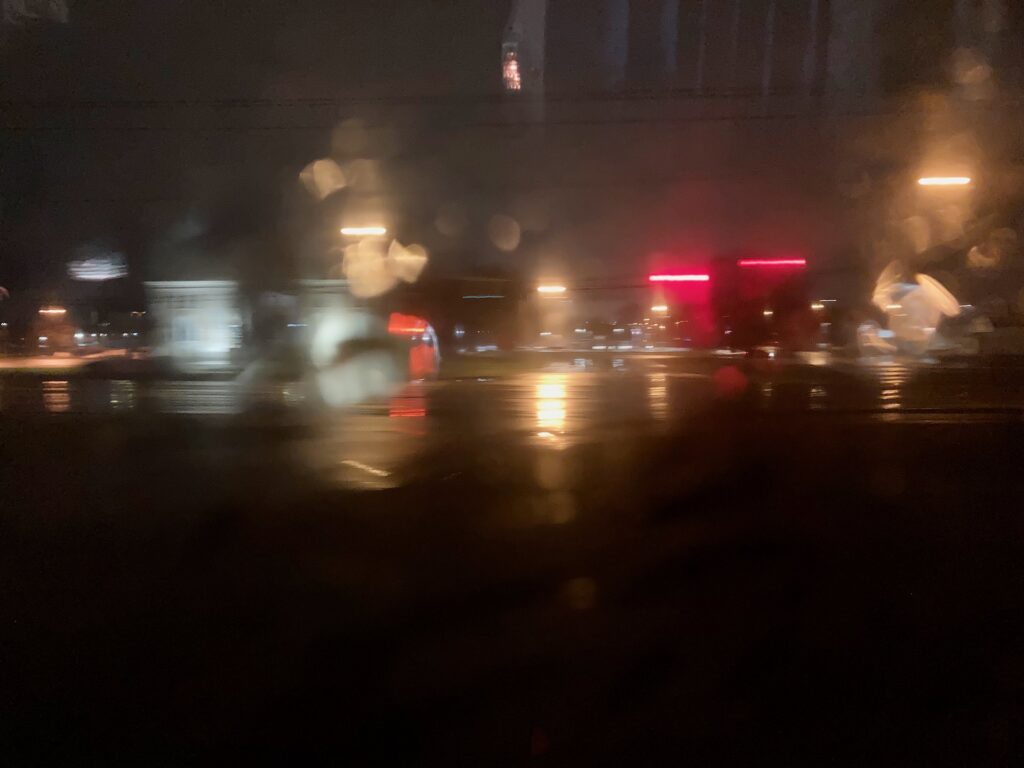 A blurred photo of a dark night, with bright lights reflecting off rain-slicked pavement.