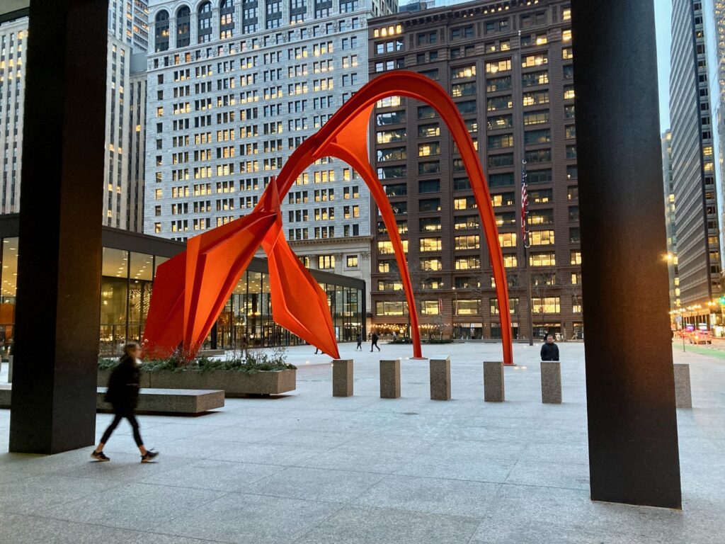 A large bright red abstract sculpture standing in a plaza surrounded by skyscrapers.