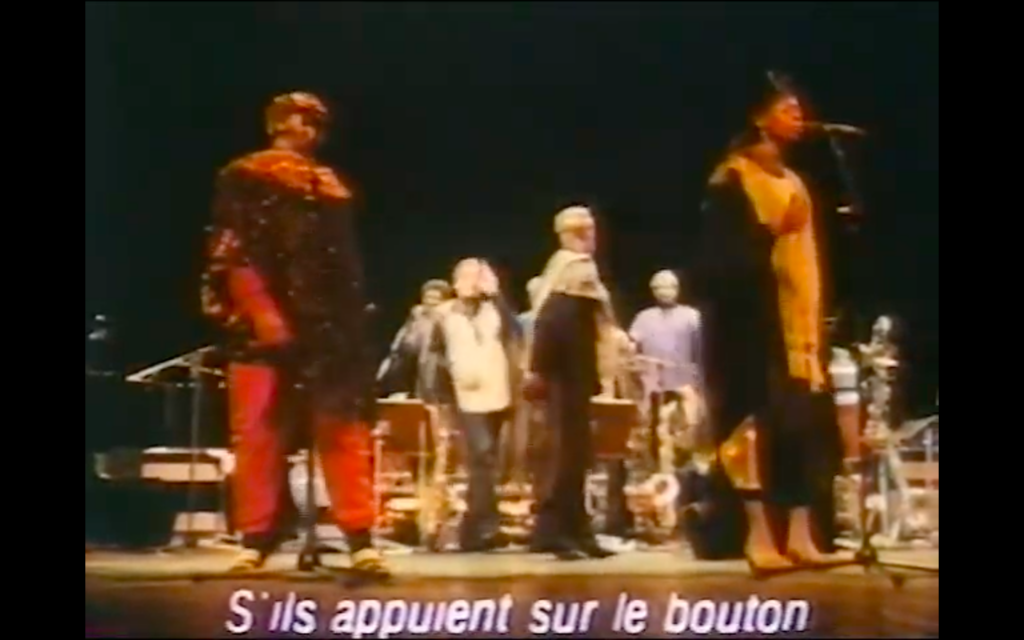 A screen grab from the 1984 film showing Sun Ra and his band, dressed in elaborate costumes, performing "Nuclear War." A subtitle in French reads, "S'ils appuient sur le bouton," i.e., "If they press the button...."
