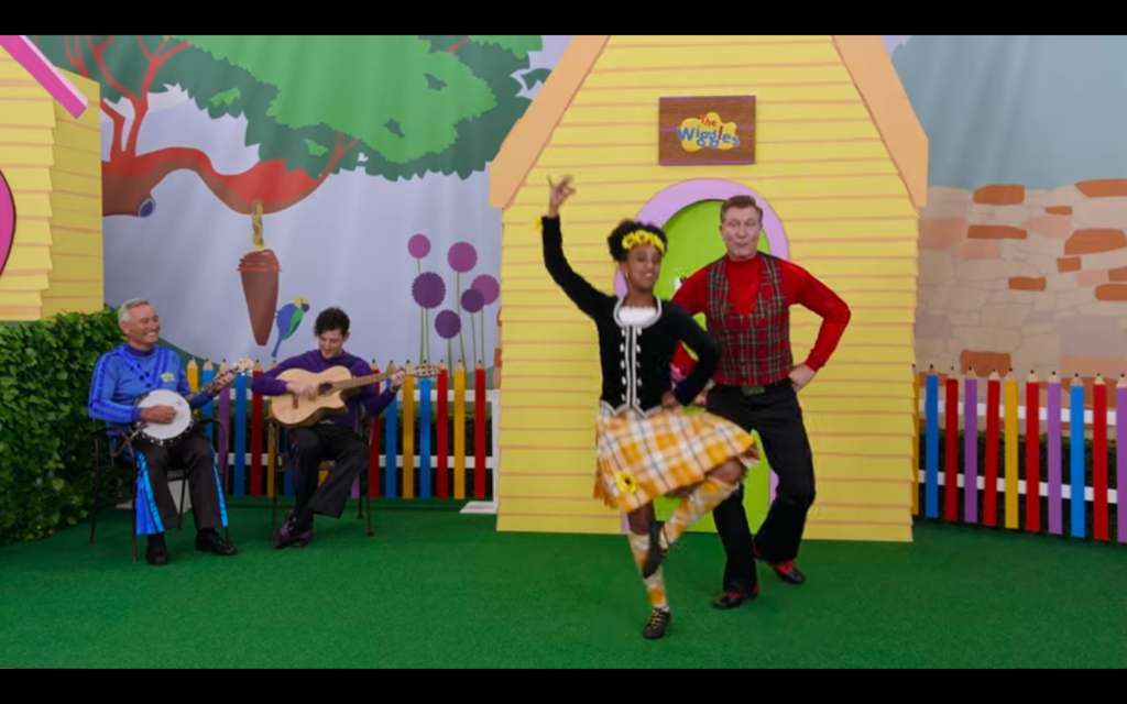 A screen grab from the video, showing a young woman wearing yellow and an older man wearing red dancing, while off to one side a man wearing blue and a man wearing purple play musical isntruments.