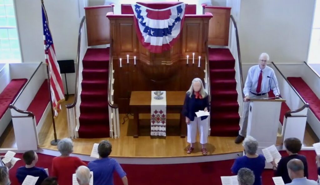 Two people standing in front of the high pulpit of the Cohasset Meeting House, with others in pews in front of them, all singing.