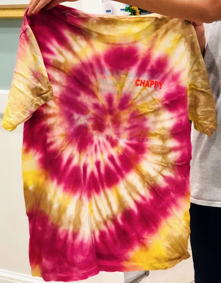Person holding a t-shirt dyed in red, yllow, and brown in a spiral pattern.