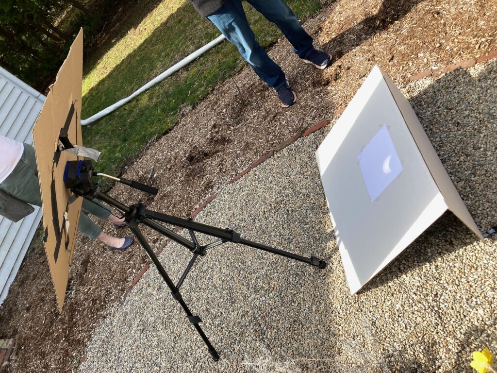 Binoculars taped to a tripod with gaffer's tape, with a large shade collar attached.The binoculars are projecting an image of the sun on a white sheet on the ground.