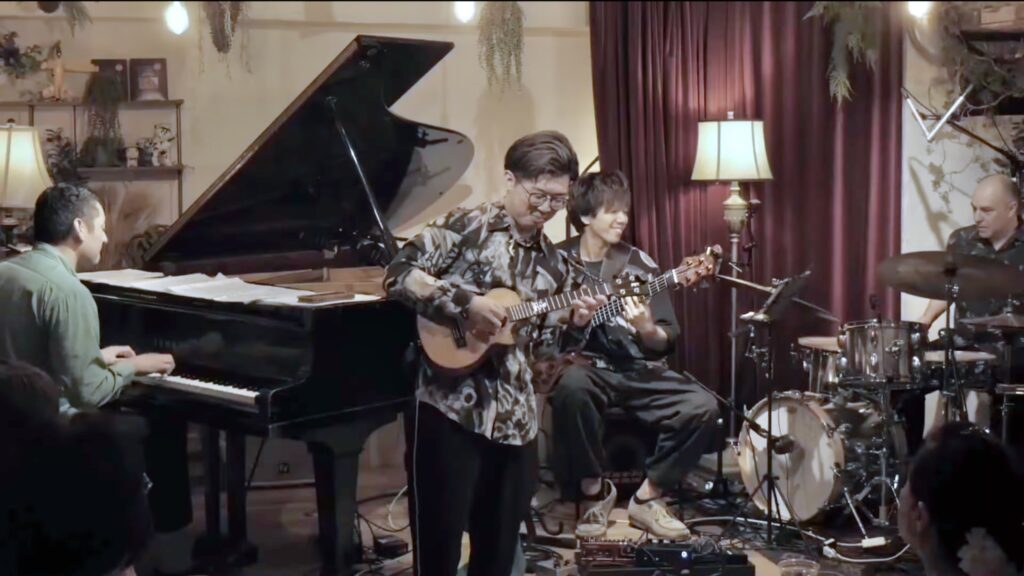Rio Saito playing ukulele, with a pianist, bassist, and drummer behind him on a small stage.