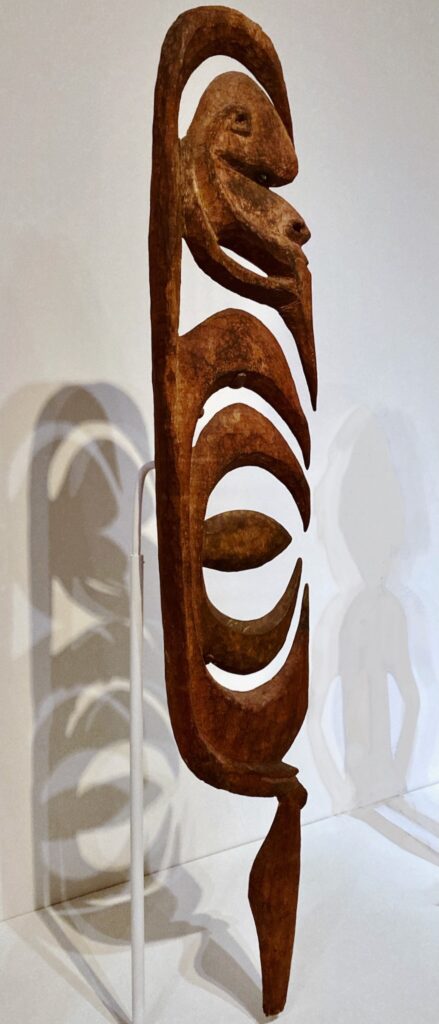 A human-sized wood sculpture with a stylized head over stylized hooks, standing on a single leg.